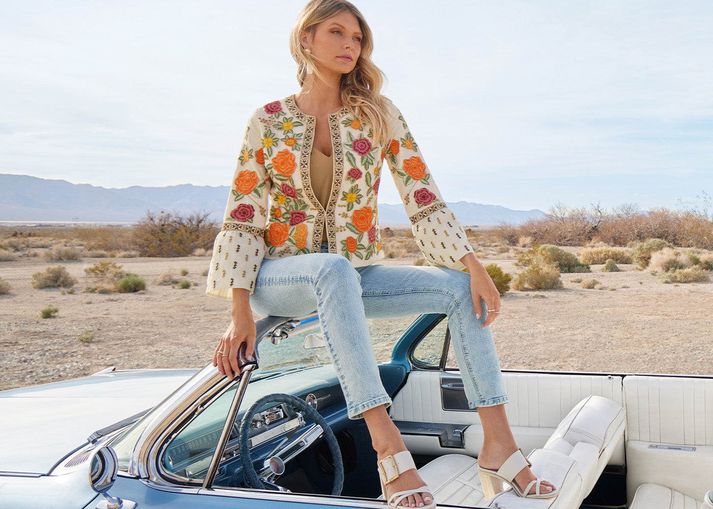 Model wearing a floral embroidered flare-sleeve jacket, tan v-neck charmeuse blouse, light wash jeans, and white buckle heels while sitting on a vintage convertible.