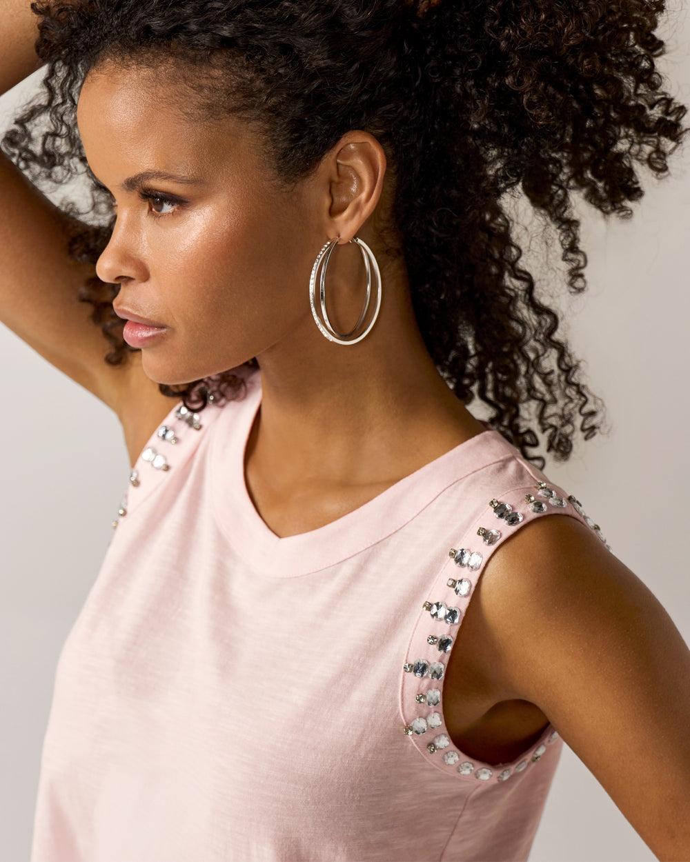 Woman wearing a pink top with embellishments and hoop earrings against a neutral background