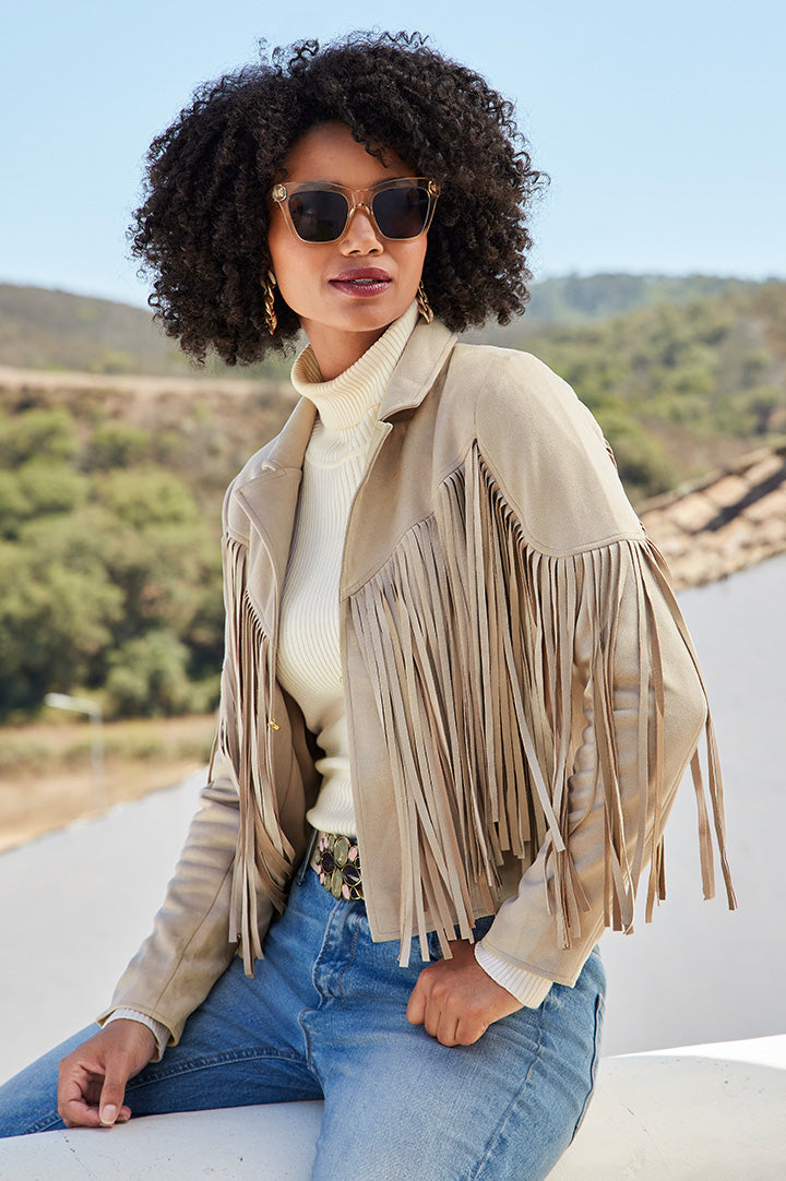 model wearing a beige fringe faux-suede jacket, off-white turtleneck sweater, jeans, and sunglasses.
