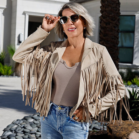 sheryl wearing a beige fringe faux suede jacket over a beige notched tank top, jeans, and sunglasses.