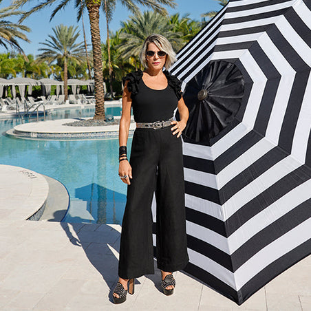 sheryl wearing a black ruffle sleeve top, embellished silver belt, black pants, and black embellished pumps standing next to a large black and white striped umbrella.