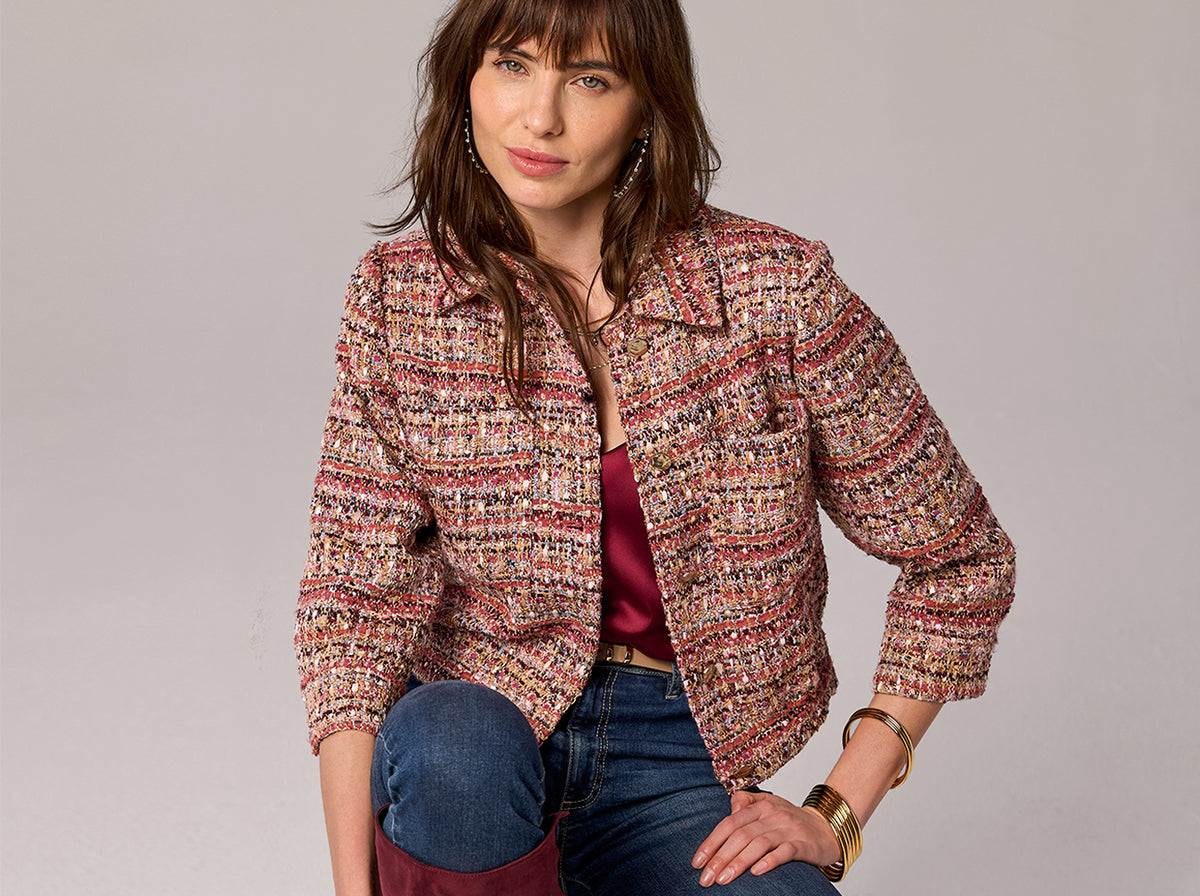 A woman models a cropped, long-sleeve tweed jacket with a multicolor pattern of pink, red, beige, and brown. She is wearing blue jeans and a burgundy top underneath, posed against a solid light gray studio background.