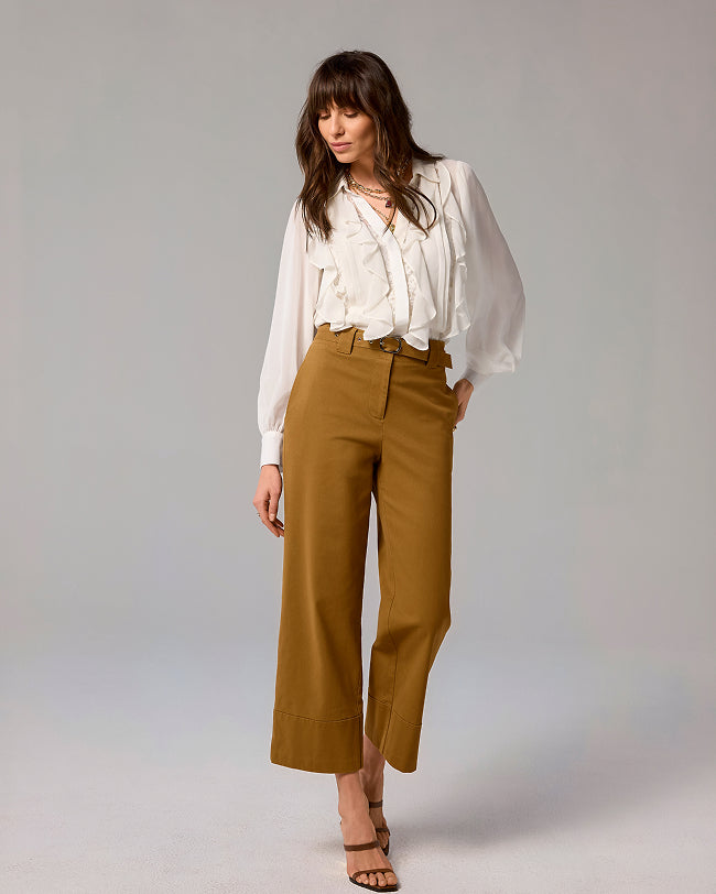 A woman modeling a white long-sleeve blouse and high-waisted, wide-leg cropped trousers in a warm camel brown, photographed against a neutral light gray studio background.