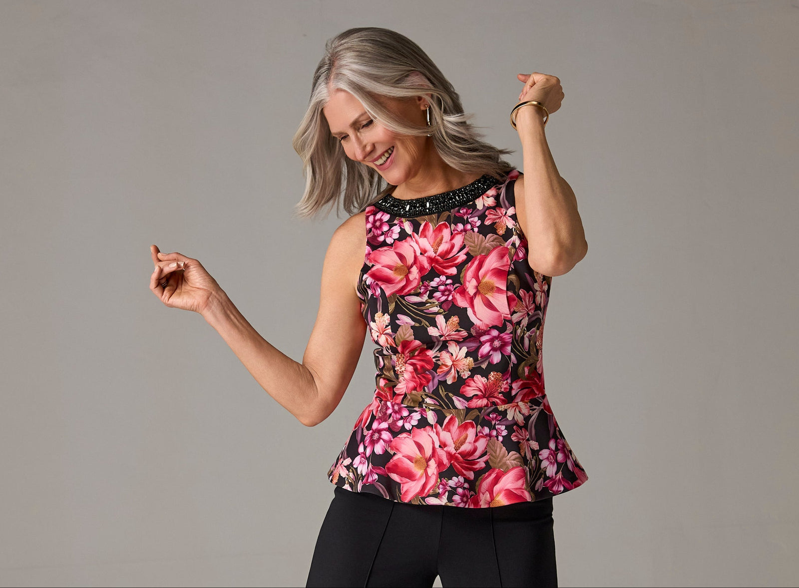 A smiling woman with gray hair models a sleeveless peplum top featuring a vibrant pink, red, and green floral pattern with a black beaded neckline, paired with black pants, against a solid gray studio background.
