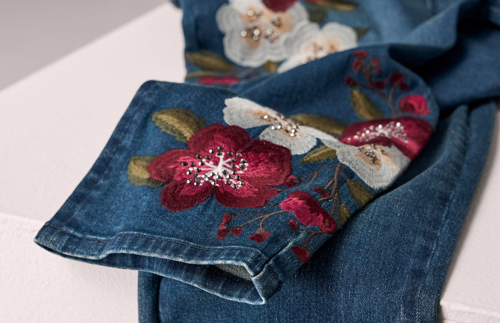 A close-up of a blue denim garment, likely a jacket or coat sleeve, featuring detailed floral embroidery in red, white, and green with sparkling rhinestone accents.
