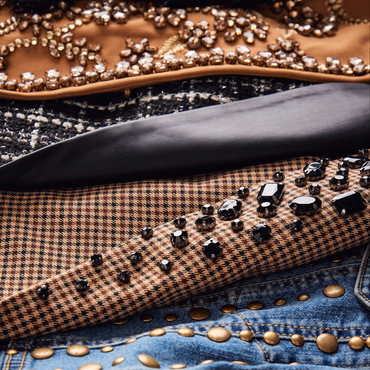 A close-up of a fashion accessory featuring a brown and black houndstooth pattern base, heavily embellished with large black faceted crystals and gold studs, layered over a black satin trim and a blue denim section with gold studs.