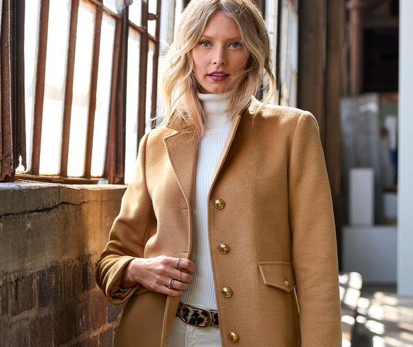 Model wearing a camel topper jacket, white ribbed turtleneck sweater, animal print belt, and white pants.