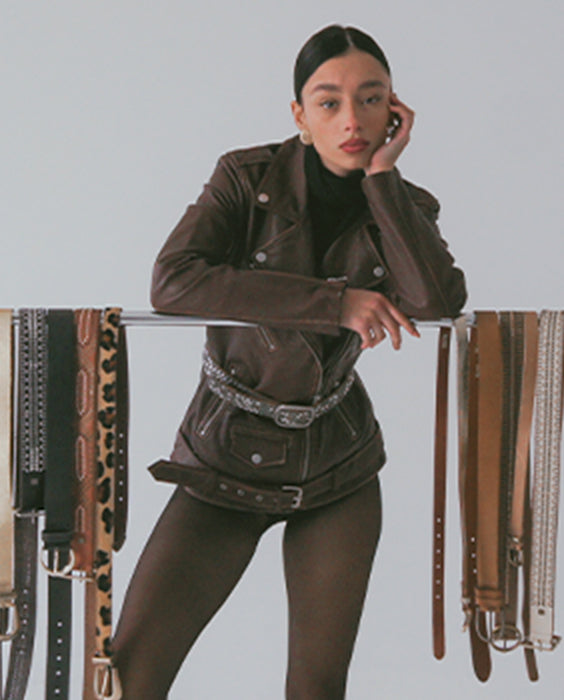 A woman wearing a dark brown faux-leather moto jacket with silver hardware and a matching belt, styled with dark brown tights. She is leaning on a metal stand decorated with various leather and faux-leather belts, set against a solid light gray background.