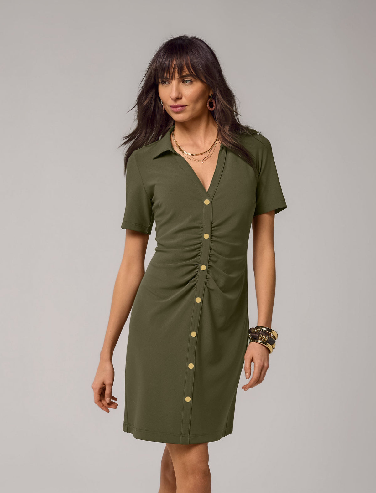 A woman modeling an olive green short-sleeve button-front dress with a gathered waist and collar, photographed against a plain light gray studio background.