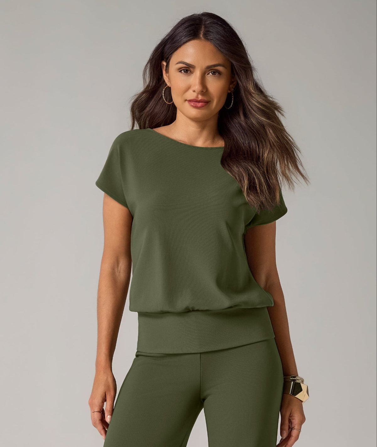 A woman modeling a matching olive green two-piece set consisting of a short-sleeve top and wide-leg pants, paired with black strappy sandals, against a neutral gray studio background.