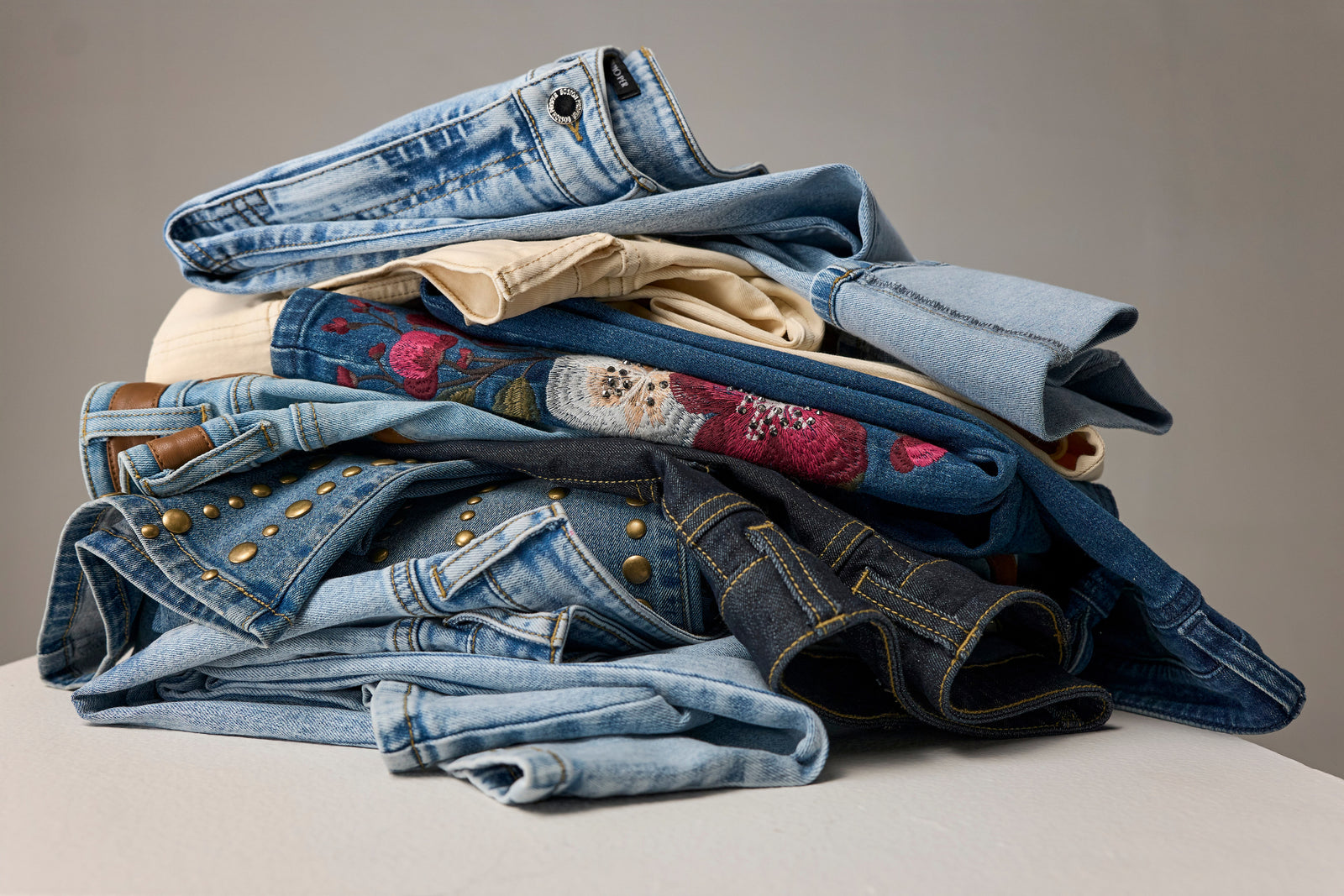 A stack of folded denim garments and jeans in various shades of blue, including light wash, dark wash, and one with colorful floral embroidery, displayed on a neutral studio background.