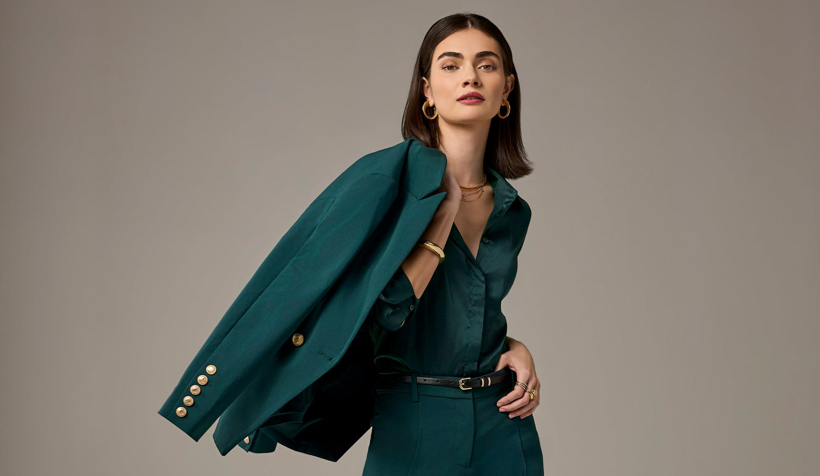 A woman modeling a dark teal green tailored blazer with gold buttons over a matching green top and skirt, posed against a solid taupe-gray studio background.