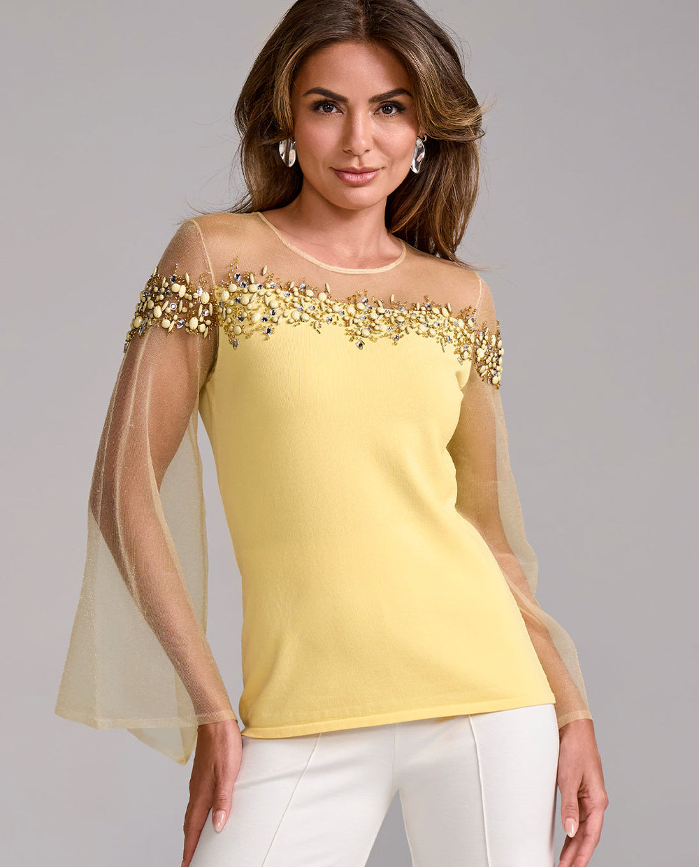 Woman wearing a yellow top with gold embellishments and sheer sleeves against a gray background