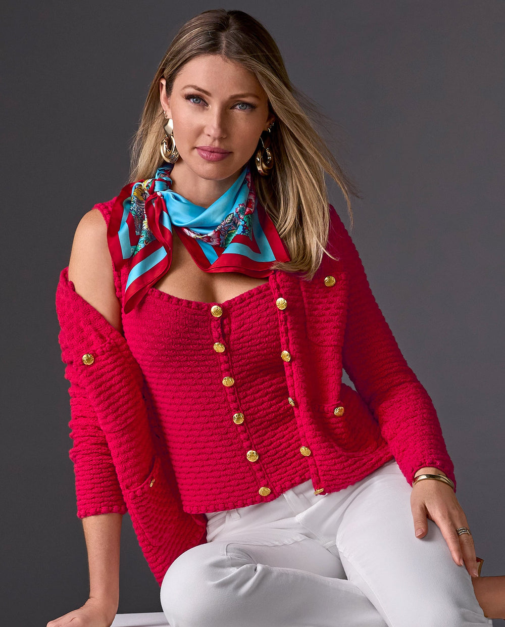 Model is wearing red cardigan and matching tank with gold buttons, blue and red scarf, and white pants with gold statement earrings.