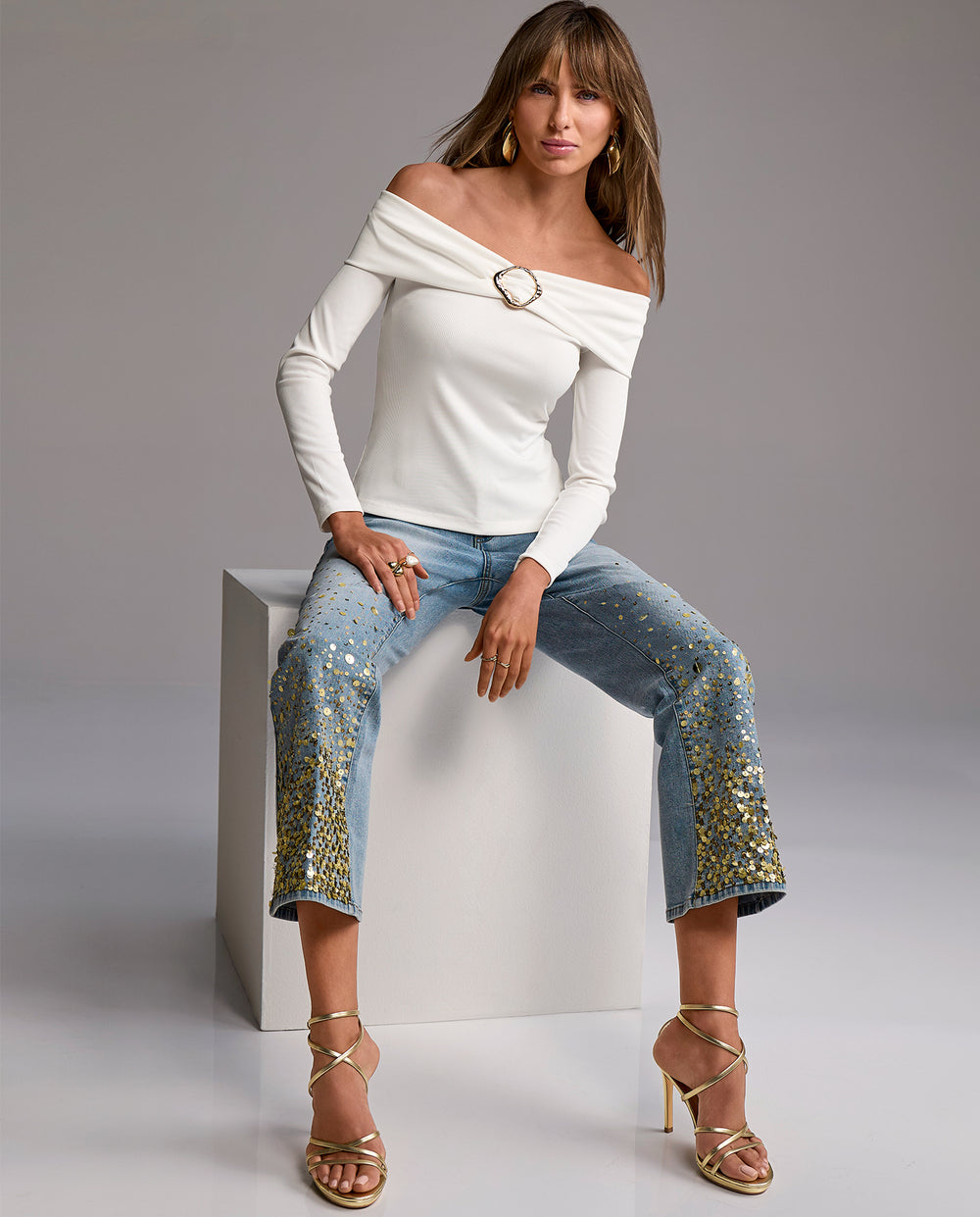 Woman sitting on a white cube wearing a white off-shoulder top and blue jeans with gold accents.
