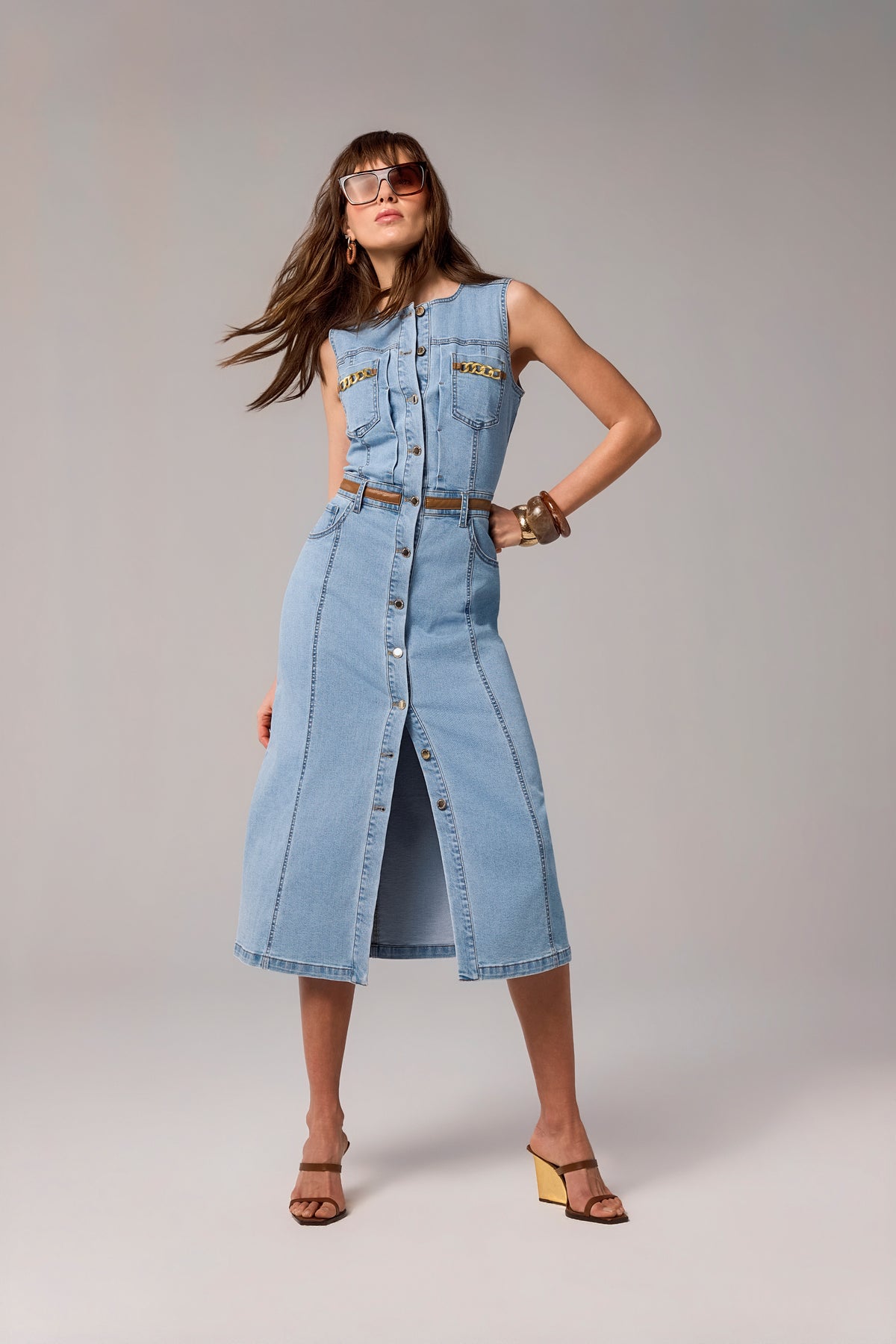 A woman modeling a light blue denim midi dress with a button-front closure, chest pockets, and a matching belt, photographed against a plain light gray studio background.