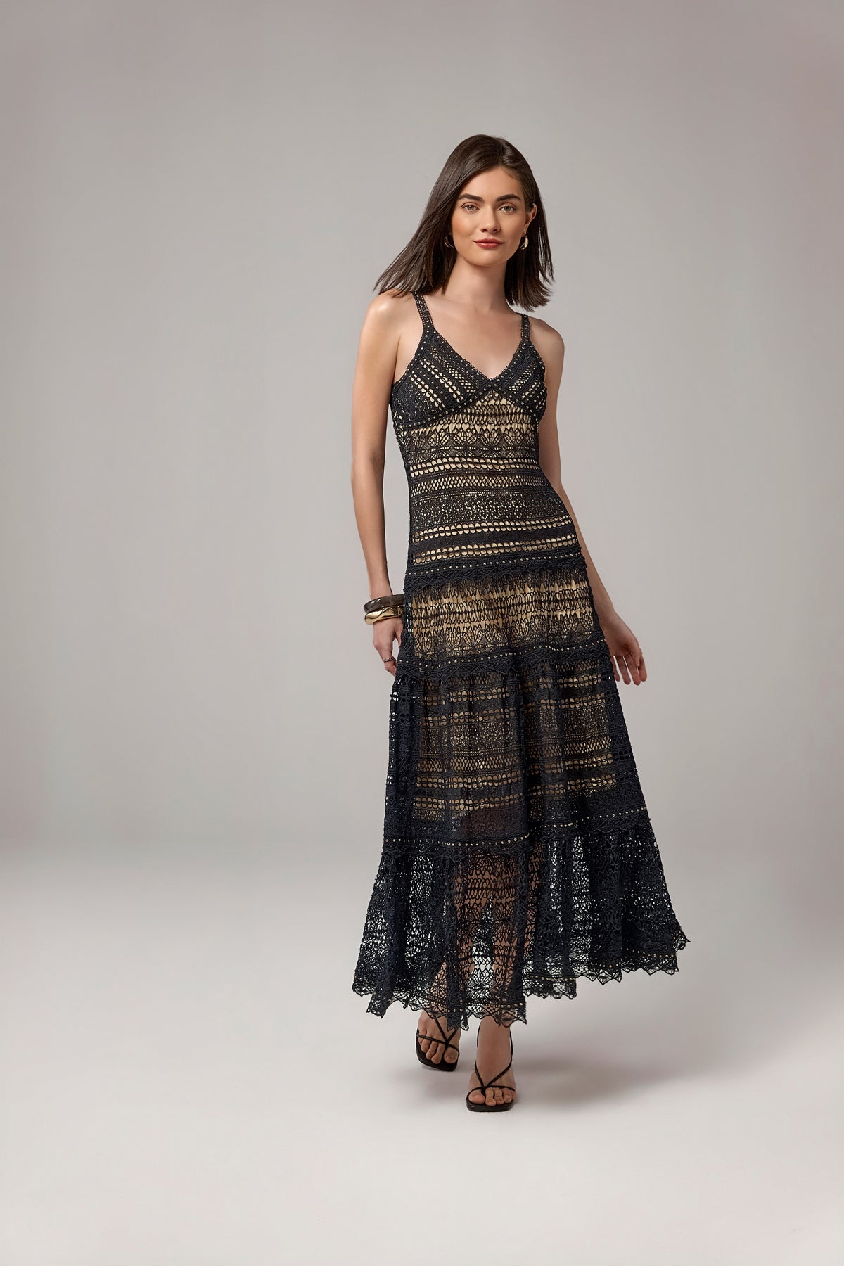 A woman modeling a sleeveless black maxi dress with a sheer, textured crochet or lace pattern and horizontal gold-toned bands, photographed against a neutral light gray studio background.