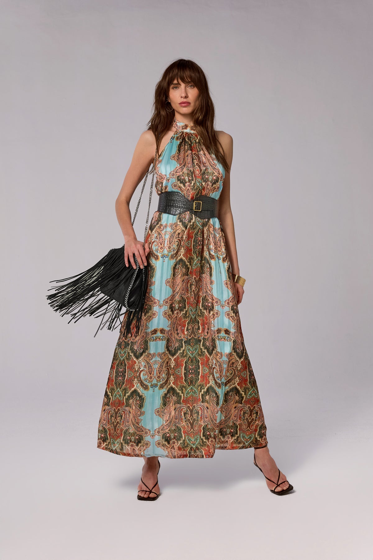A woman modeling a sleeveless halter-neck maxi dress with a colorful ornate paisley-style print in turquoise, rust, green, and beige, cinched at the waist with a wide black belt. She is holding a black fringed handbag against a plain light gray studio background.