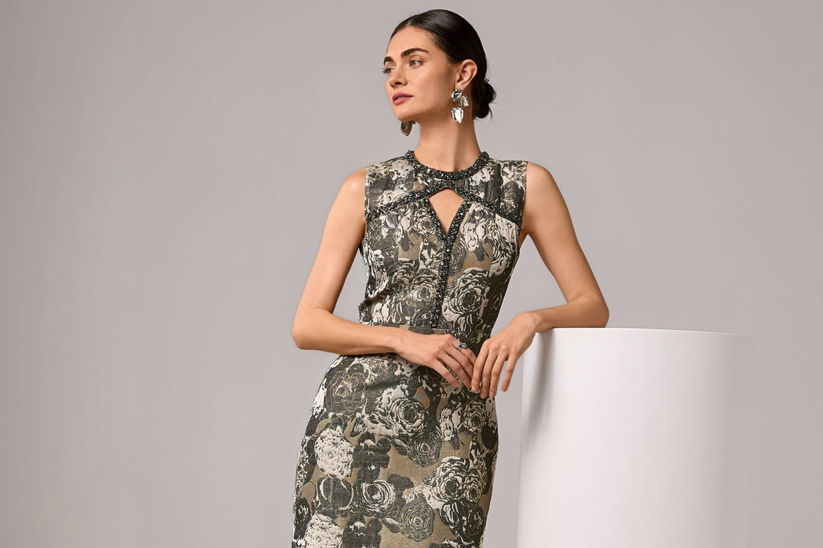 A woman models a sleeveless mini dress with a floral pattern in olive green, gray, and beige tones. She is standing next to a white cylindrical pedestal with a faint logo on a light gray studio background.