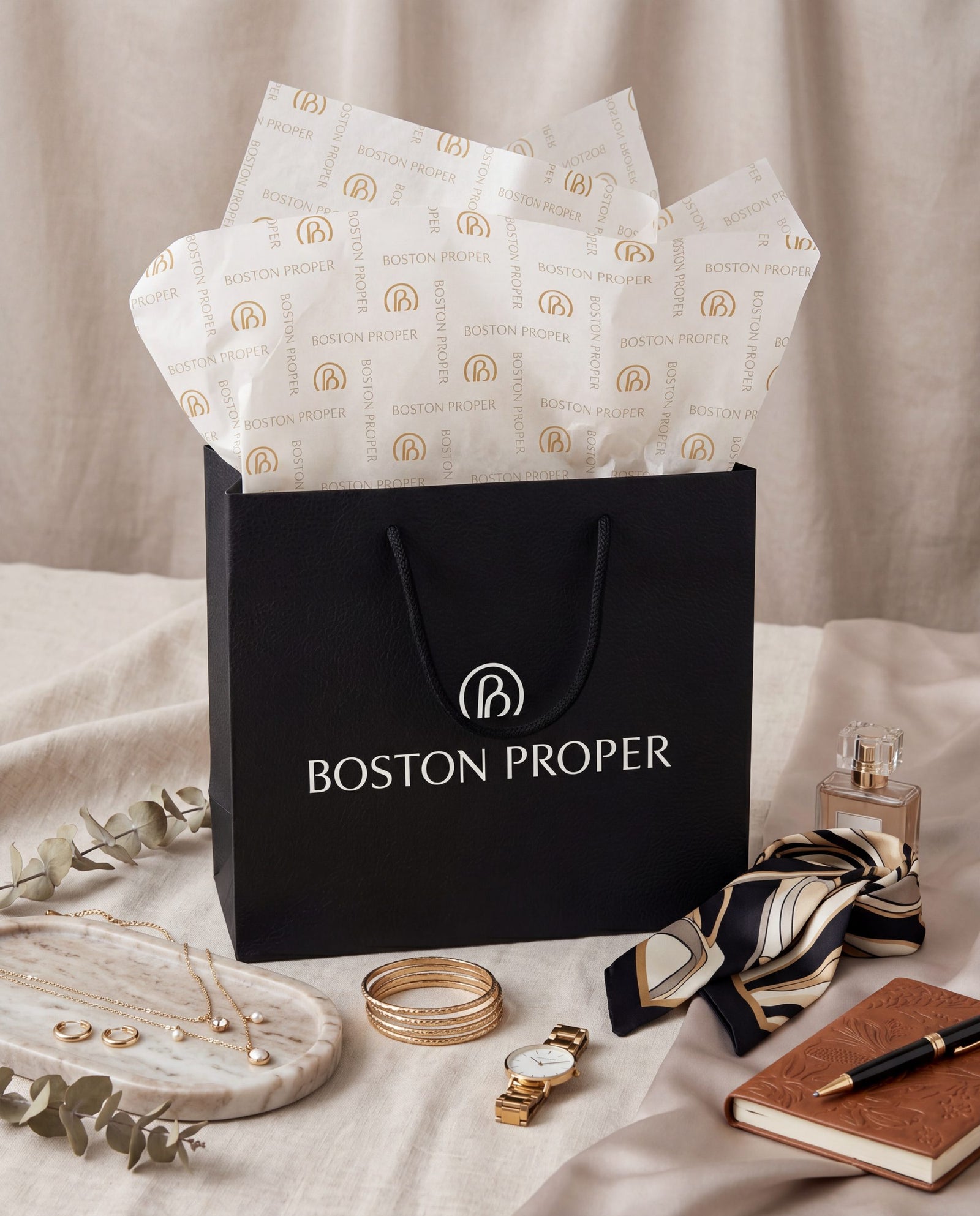 A black Boston Proper gift bag filled with branded white tissue paper, styled on draped beige fabric with jewelry, a scarf, a watch, a notebook, and a perfume bottle around it.