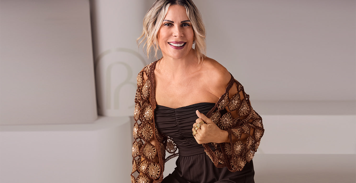 A smiling blonde woman wearing a brown off-the-shoulder dress with a sheer patterned shawl draped over one shoulder, posing in a minimalist studio setting.
