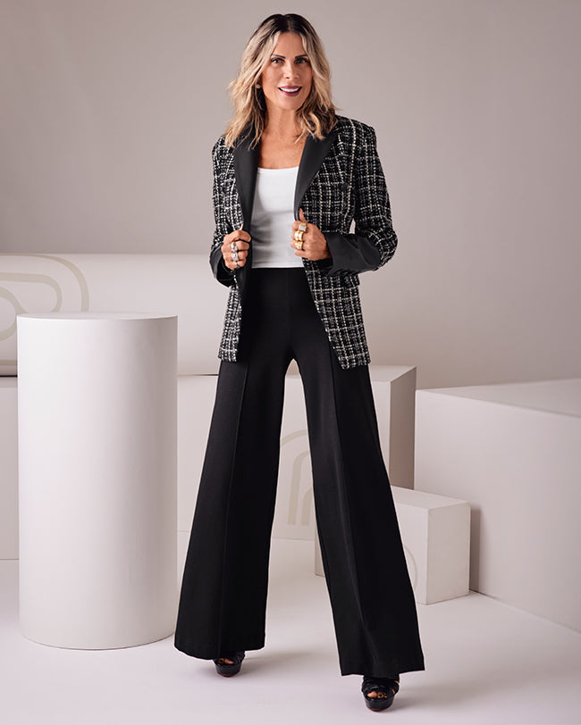 A woman models a black and white plaid blazer with black lapels, worn over a white top and black wide-leg pants, standing in a minimalist studio setting with white geometric pedestals.