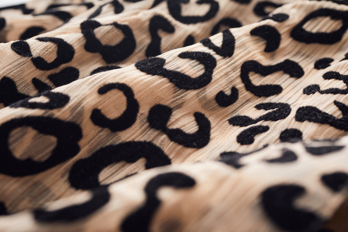 Close-up of a leopard print fabric with black irregular spots on a warm beige background, showing soft folds and texture.
