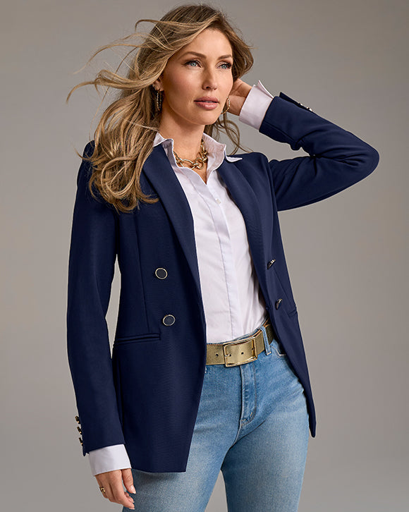 Woman wearing a navy blazer, white shirt, and blue jeans on a gray background
